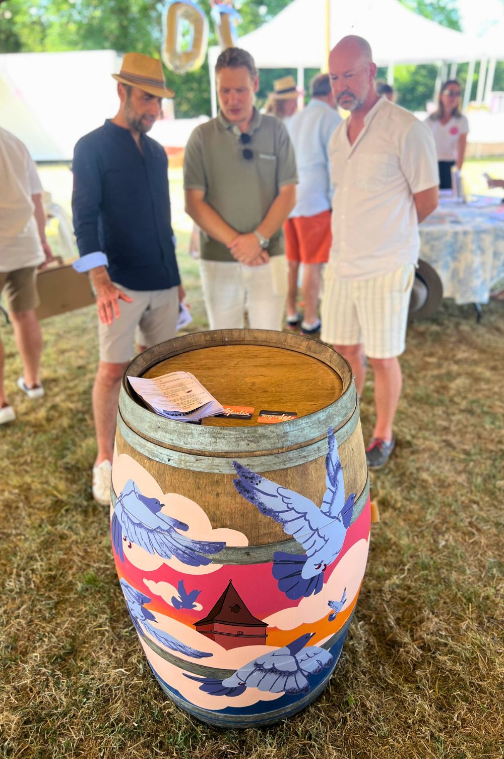 des pigeons et un pigeonnier peints sur une barrique de vin devant trois personnes. Celle du milieu est Paul Marnata, créateur des Muralistes, entouré par les acheteurs de la barrique.