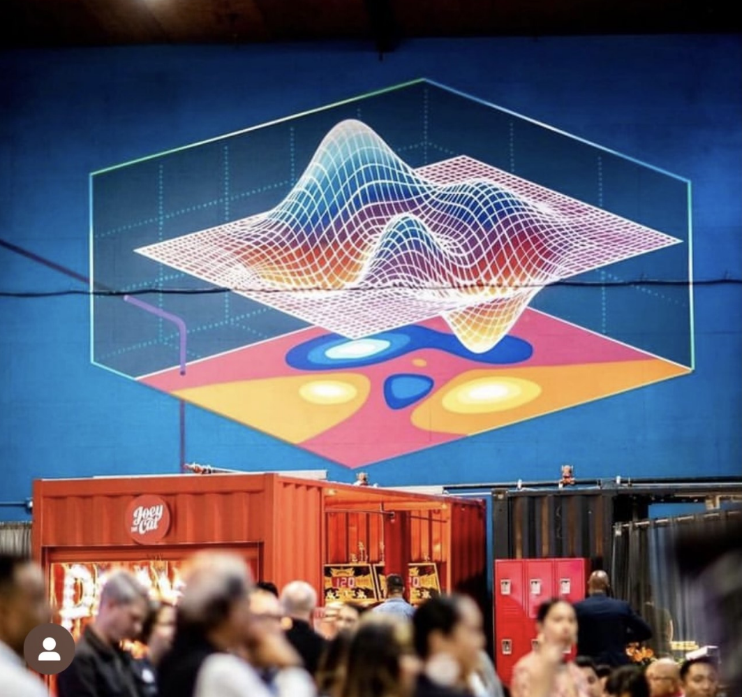 fresque murale représentant des ondes colorées en bleu, orange et rose sur fond bleu. Au bas de l'image, des personnes dans la salle.