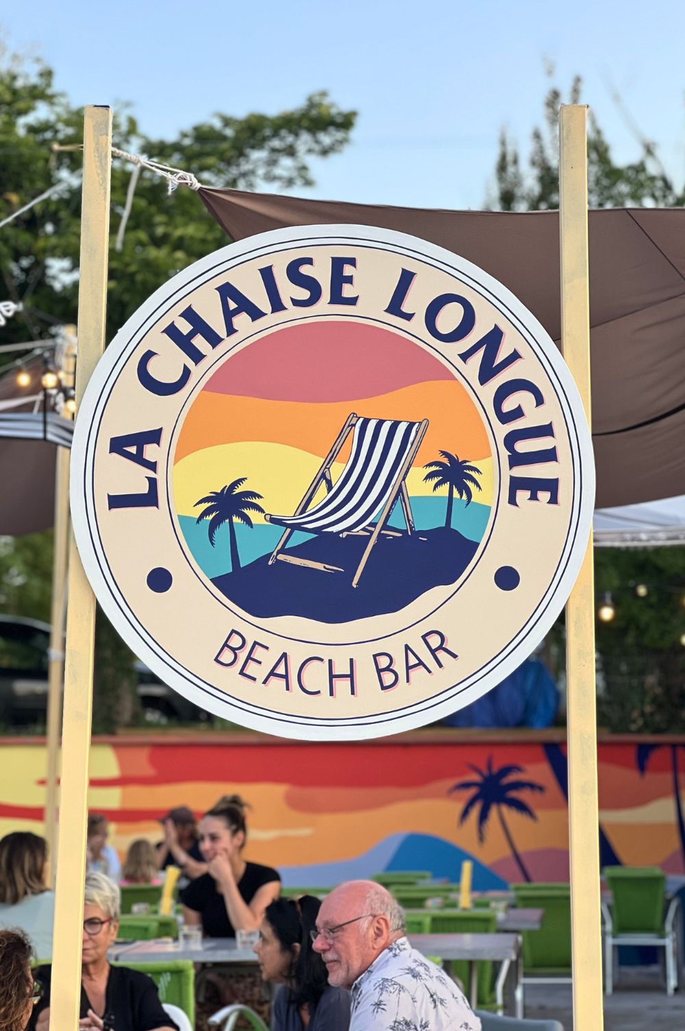enseigne ronde peinte sur un panneau de bois, avec inscrit la chaise longue en haut, beach bar en bas, et au milieu l'illustration d'un transat avec des palmiers.