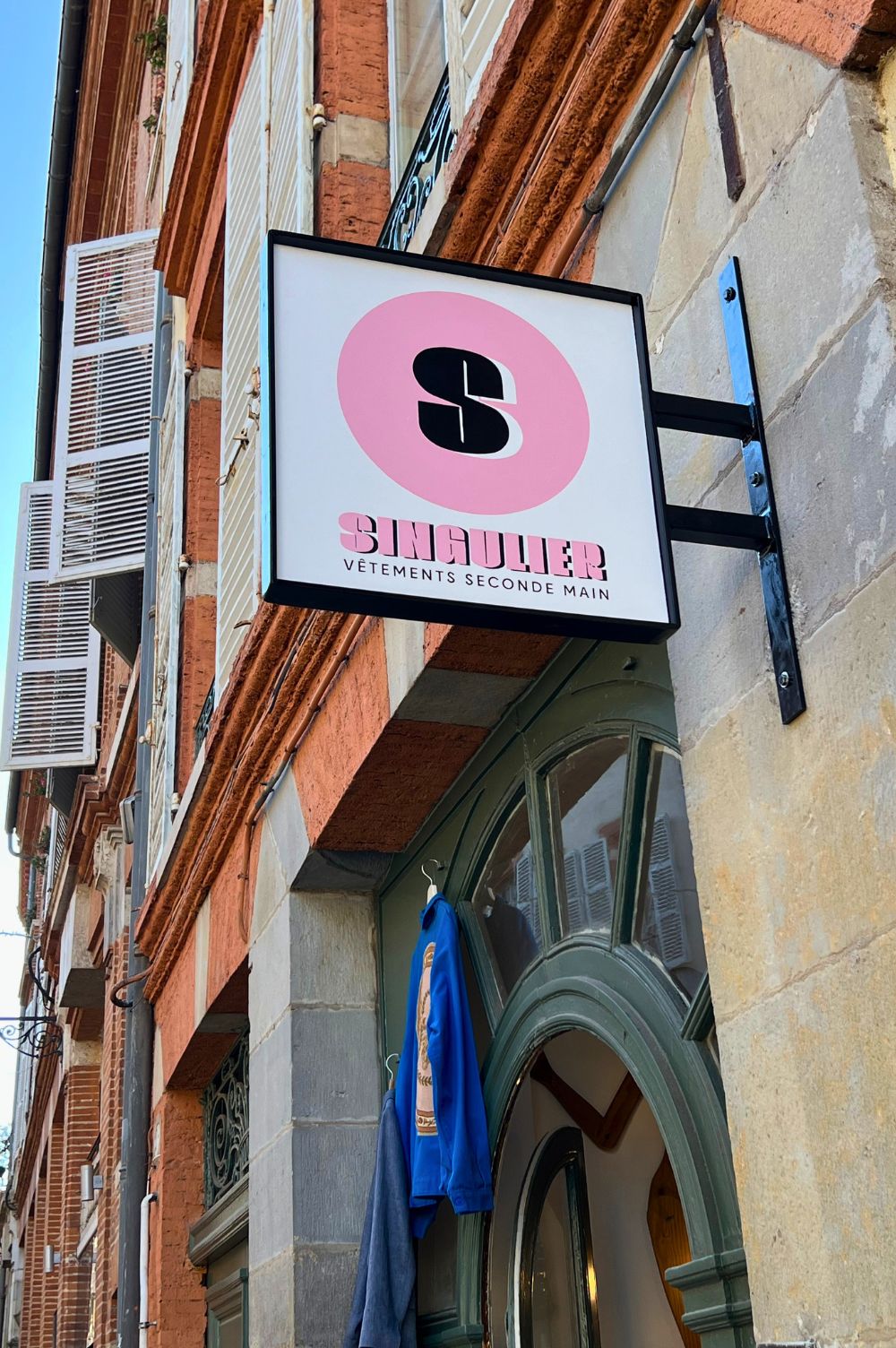 enseigne peinte à la main avec structure en métal, un S dans un cercle rose et l'inscription SINGULIER en dessous, le nom de la boutique toulousaine.