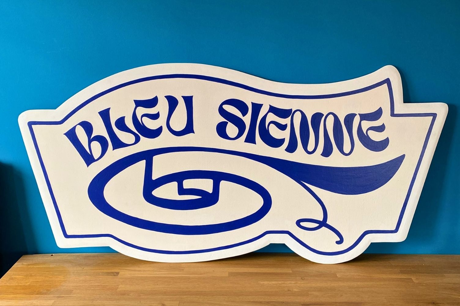 enseigne en bois peint représentant le bas d'un tapis roulé et l'inscription au-dessus BLEU SIENNE.