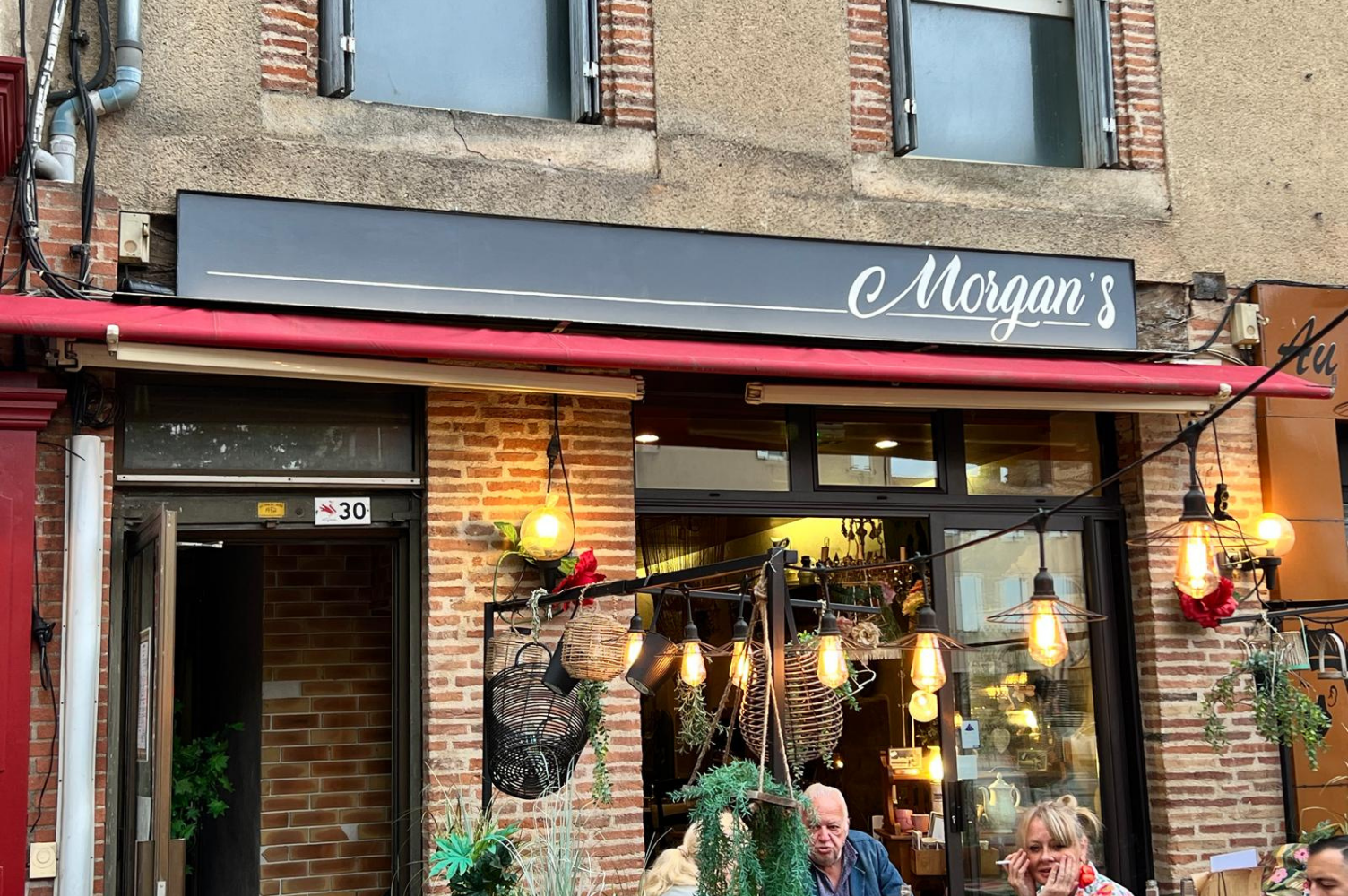 enseigne grise avec l'inscription "Morgan's" au-dessus des portes du restaurant. Sur la terrasse, quelques guirlandes et des personnes qui profitent à une table.