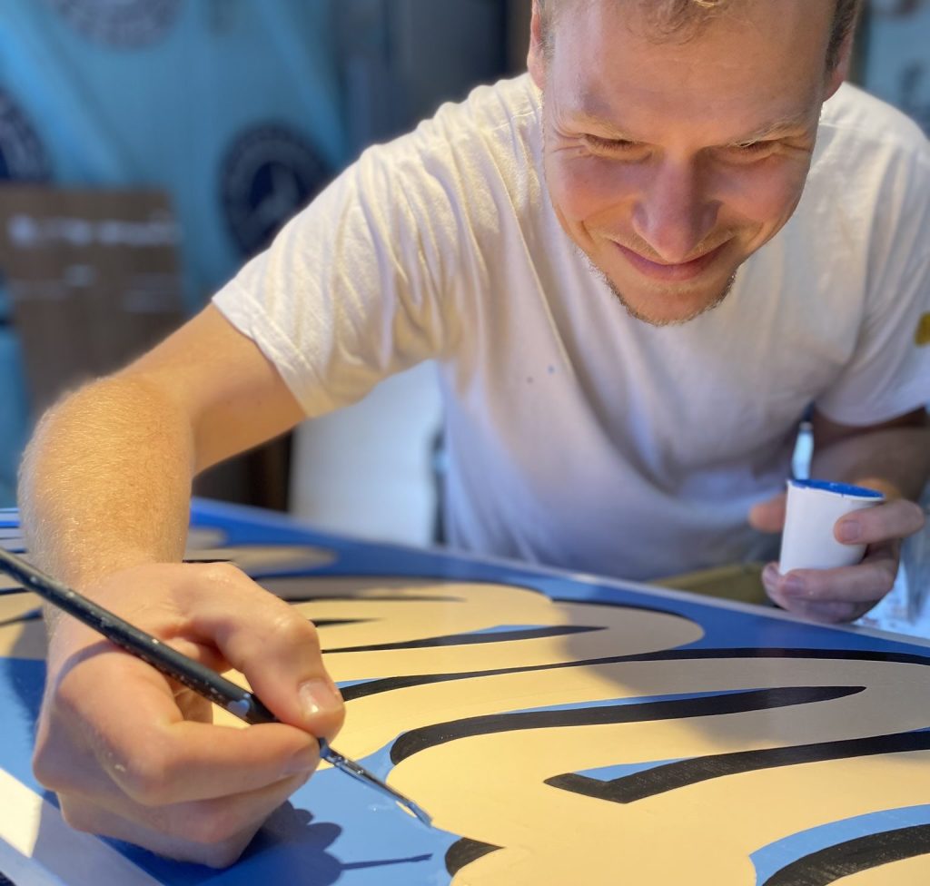 Paul Marnata est en train de peindre des lettres sur une enseigne pinceau à la main, il tient son pot de peinture dans l'autre main.
