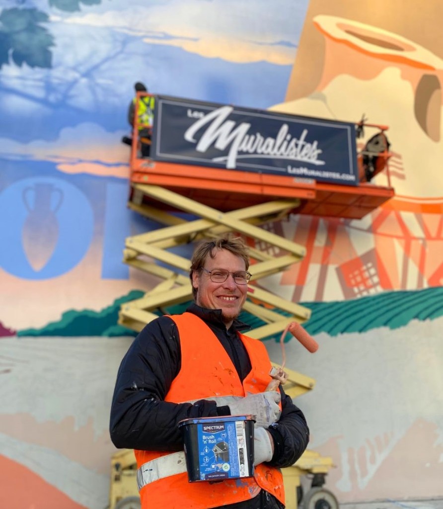 Paul Marnata, créateur des Muralistes, avec un gilet orange fluo tient un pot de peinture et un rouleau devant une grande fresque murale qu'il est en train de peindre. Sur la nacelle derrière lui se trouve un grande banderole avec l'inscription Les Muralistes.