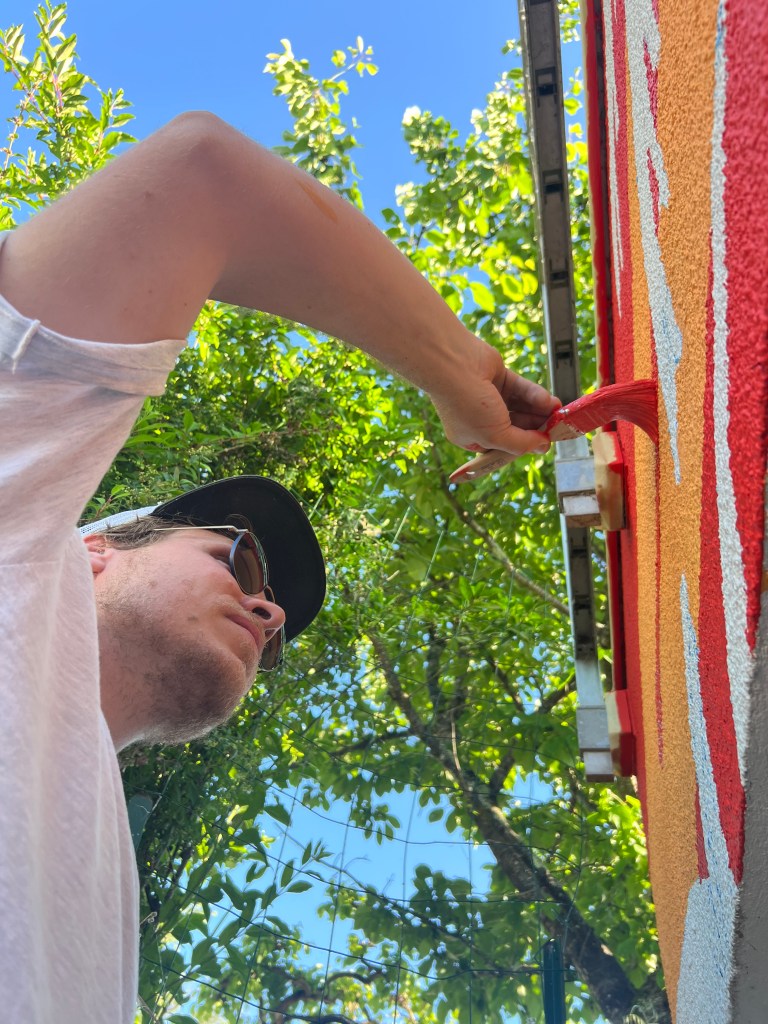 Paul Marnata des Muralistes vu du dessous, en train de peindre un mur avec un pinceau et une peinture rouge, sous des arbres et un ciel bleu.